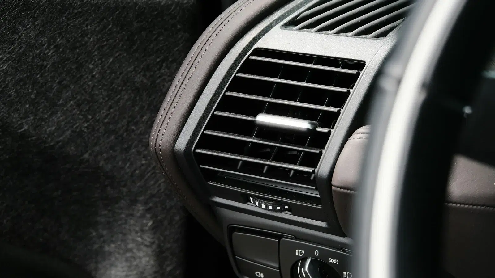 Close-up of a car dashboard air vent with a chrome adjuster tab, set in a brown leather dash
