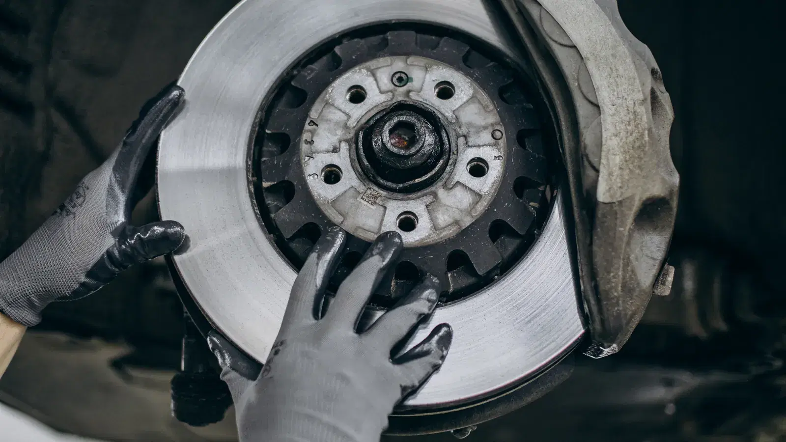 Gloved hands holding a brake disc on a car with the wheel removed, the caliper fitted over the disc
