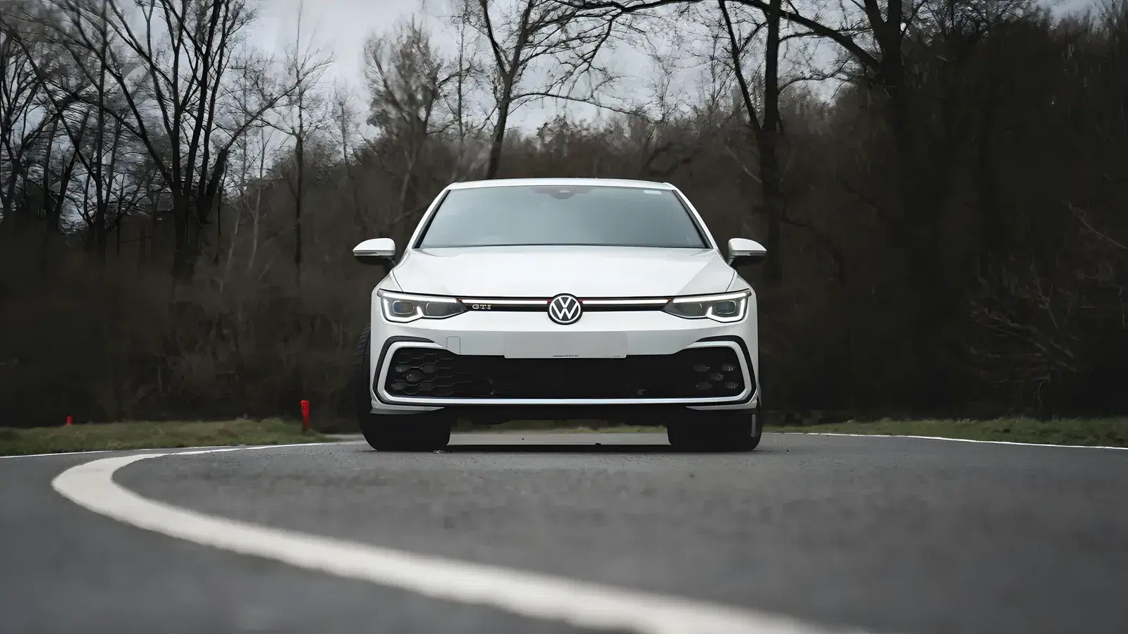 White Volkswagen Golf GTI seen head on, driving through a bend on a grey day with bare trees behind