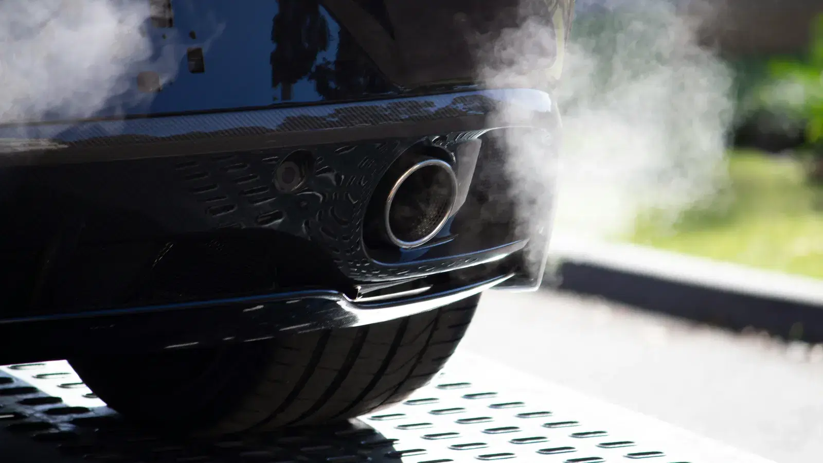 White vapour drifting from the chrome exhaust tip of a dark car, parked on a metal ramp