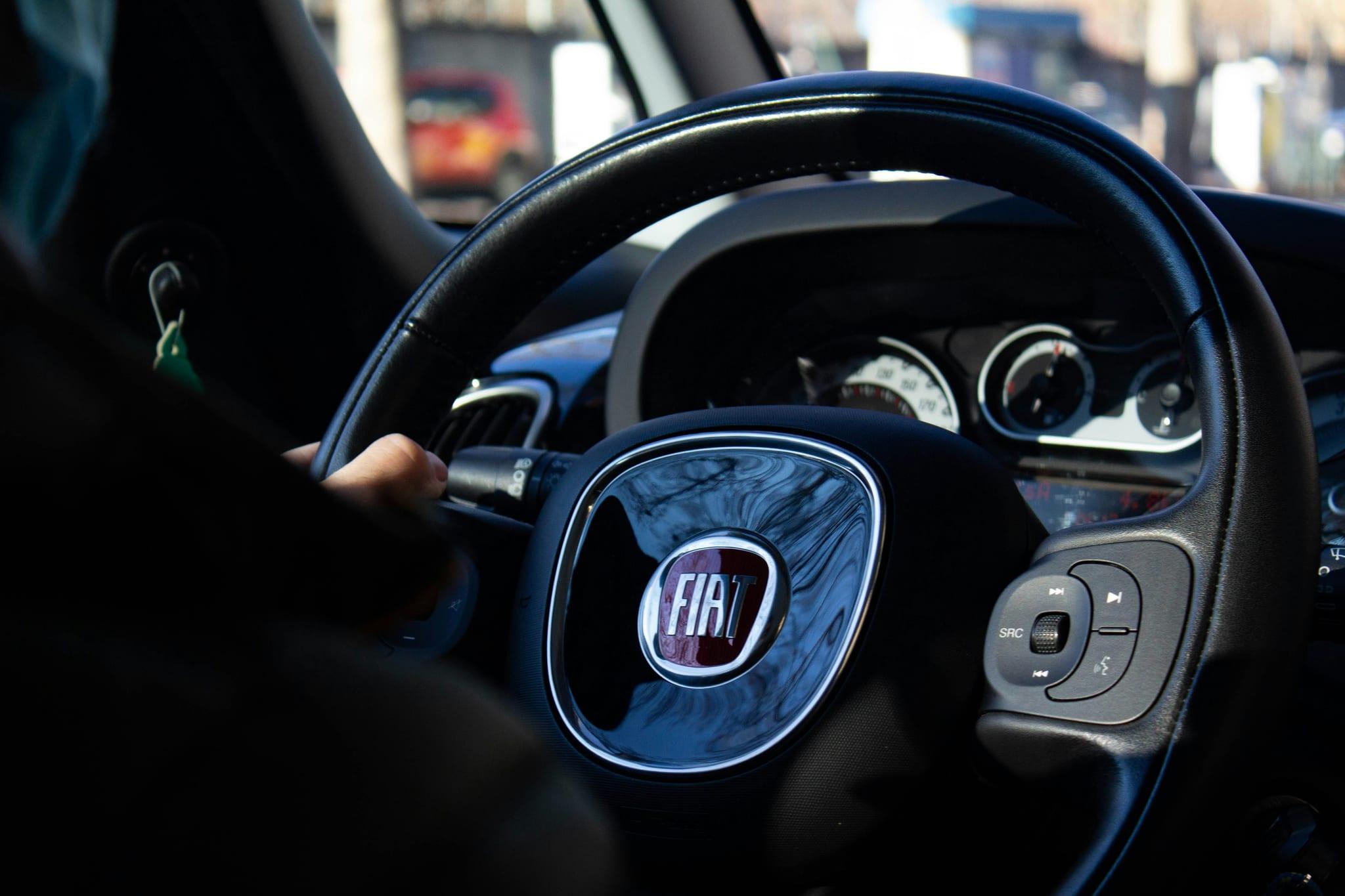 Close-up of the Fiat badge on a steering wheel, dashboard gauges beyond